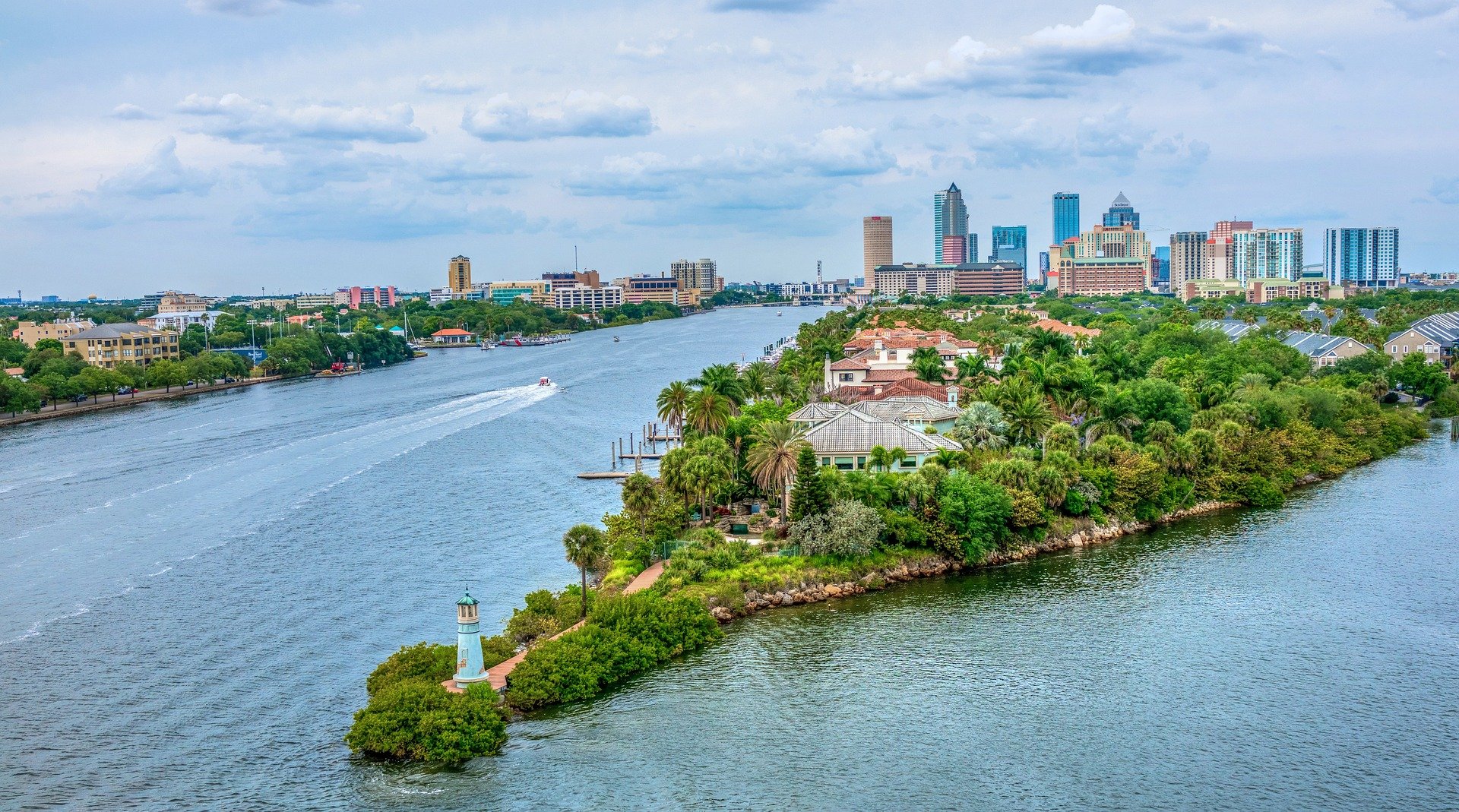 A city skyline with tall modern buildings in the background and a waterway with boats in the foreground, with residential houses and a small island with a lighthouse and lush greenery.