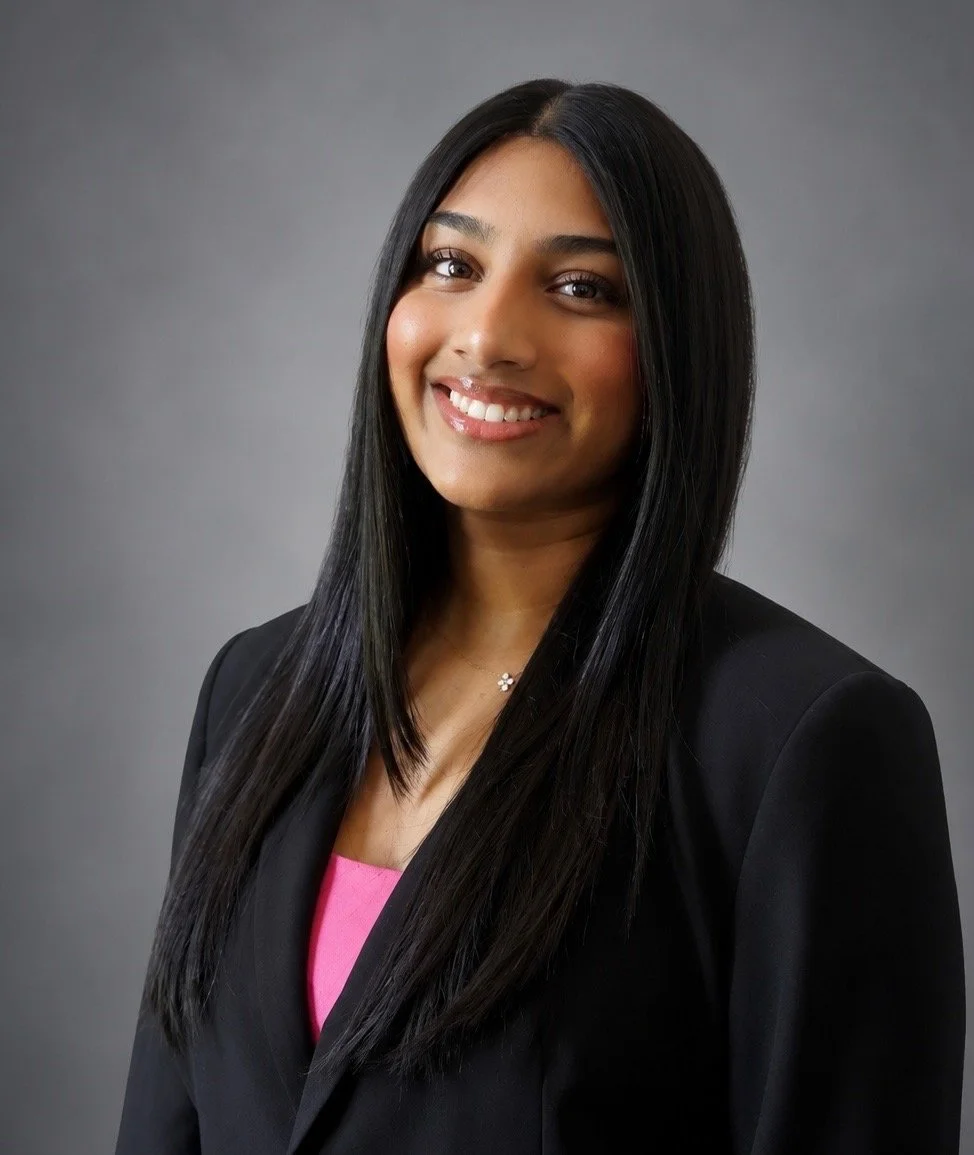 Professional woman with long black hair, smiling, wearing a black blazer and a pink top, against a gray background.