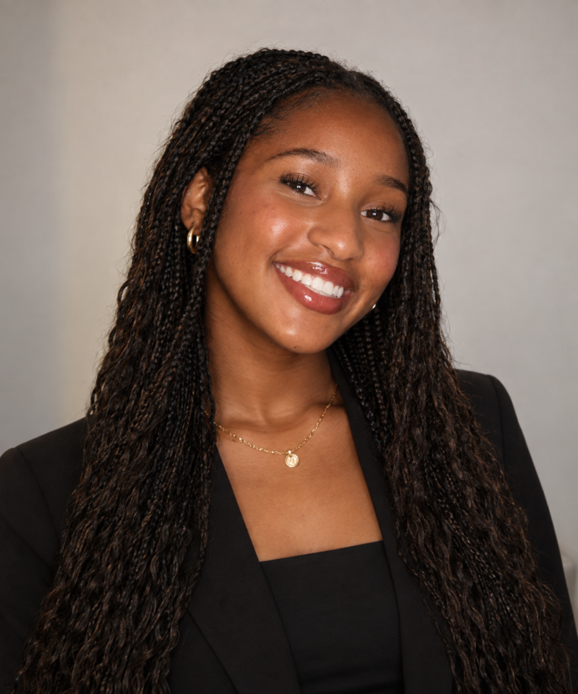 A woman with long twisted hair smiling at the camera, wearing a black blazer, gold hoop earrings, a delicate gold necklace, and a black top against a neutral background.