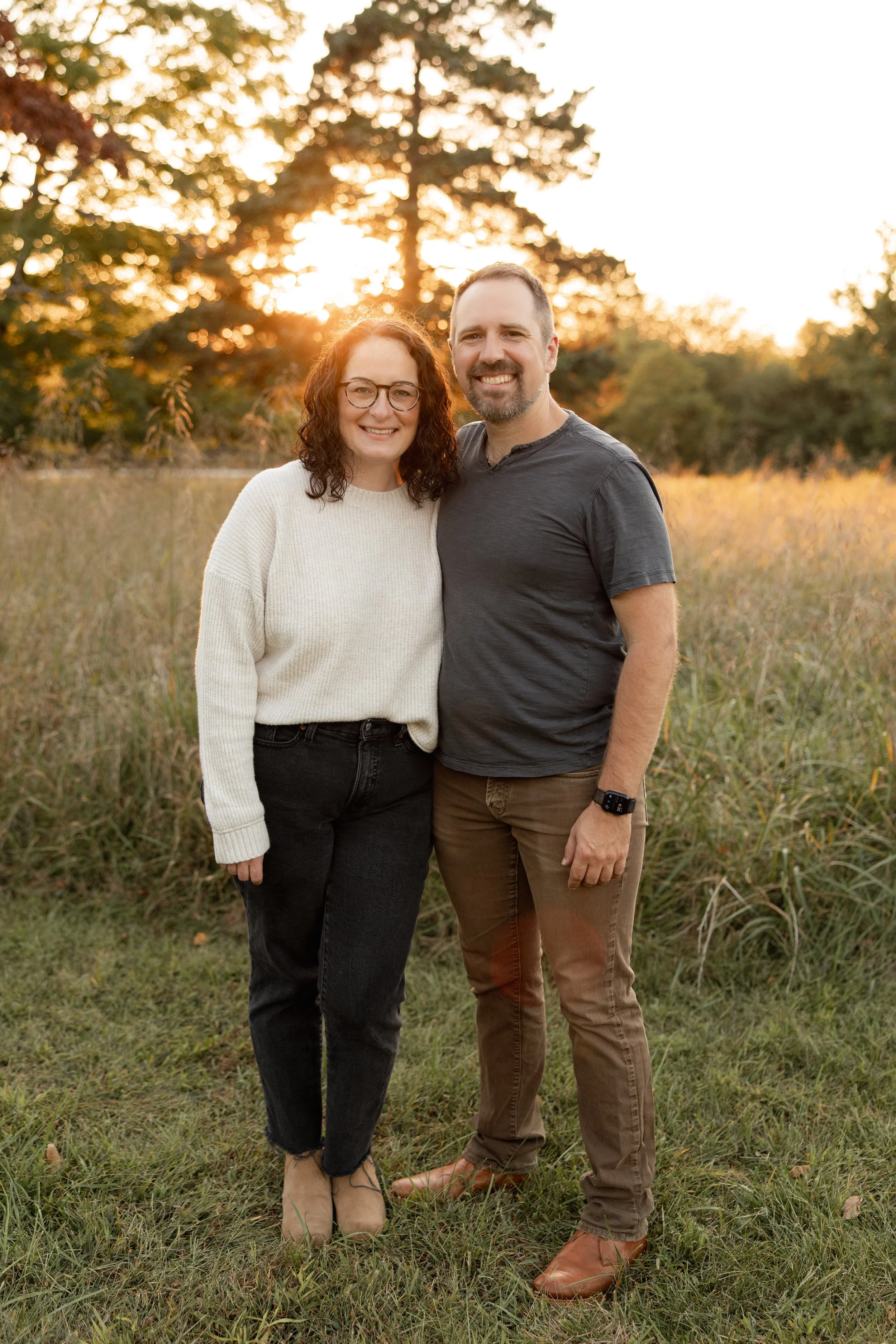 A smiling couple standing outdoors in a field during sunset. The woman has curly hair, glasses, and is wearing a white sweater and black pants. The man has short hair, a beard, and is wearing a dark gray t-shirt and brown pants.