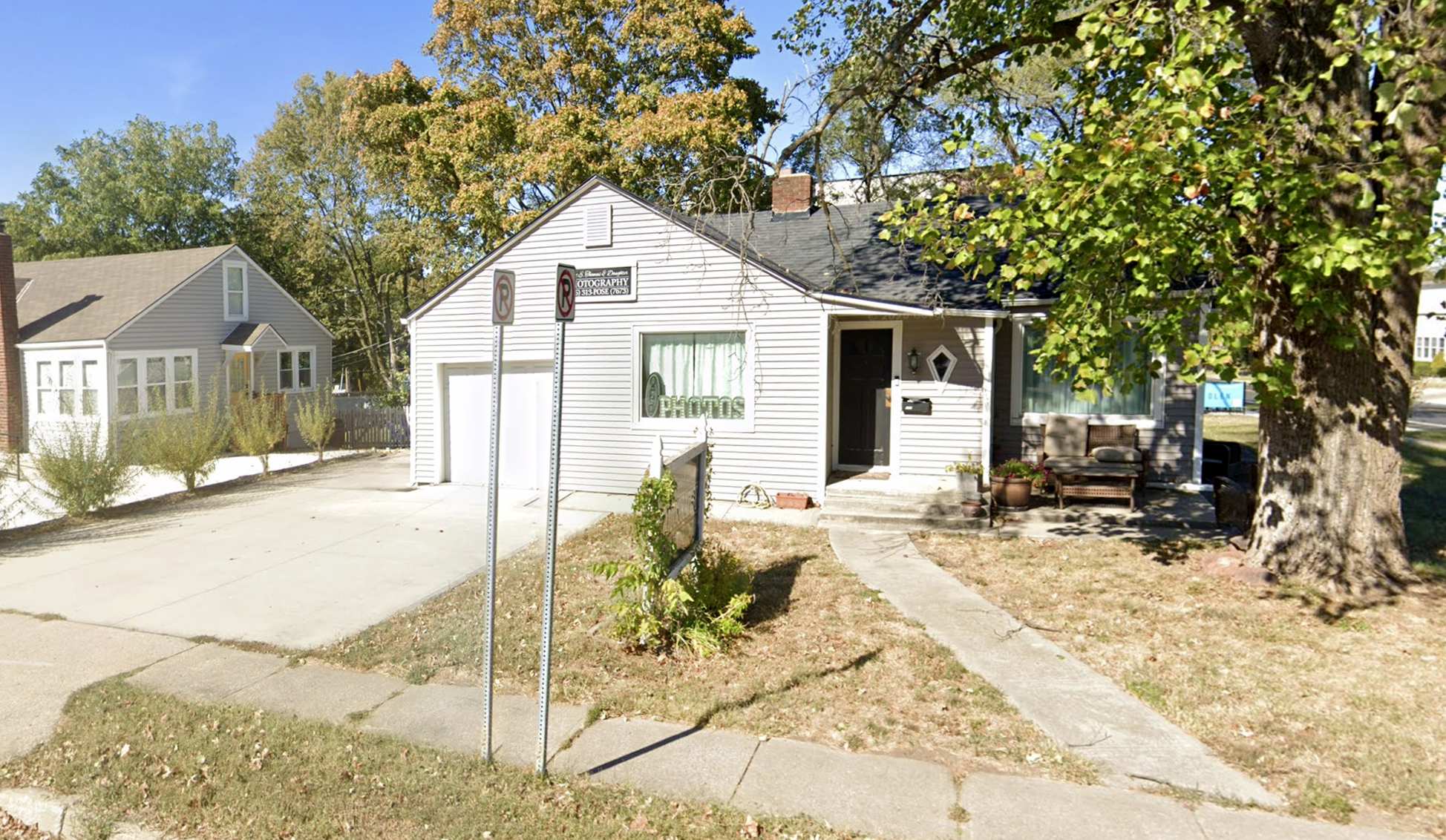 White house with a walkway leading to the front door, large trees with green leaves, and a small sign that reads 'PHOTOS' in the window. Two no-parking signs are in front of the house. There are chairs and potted plants outside.
