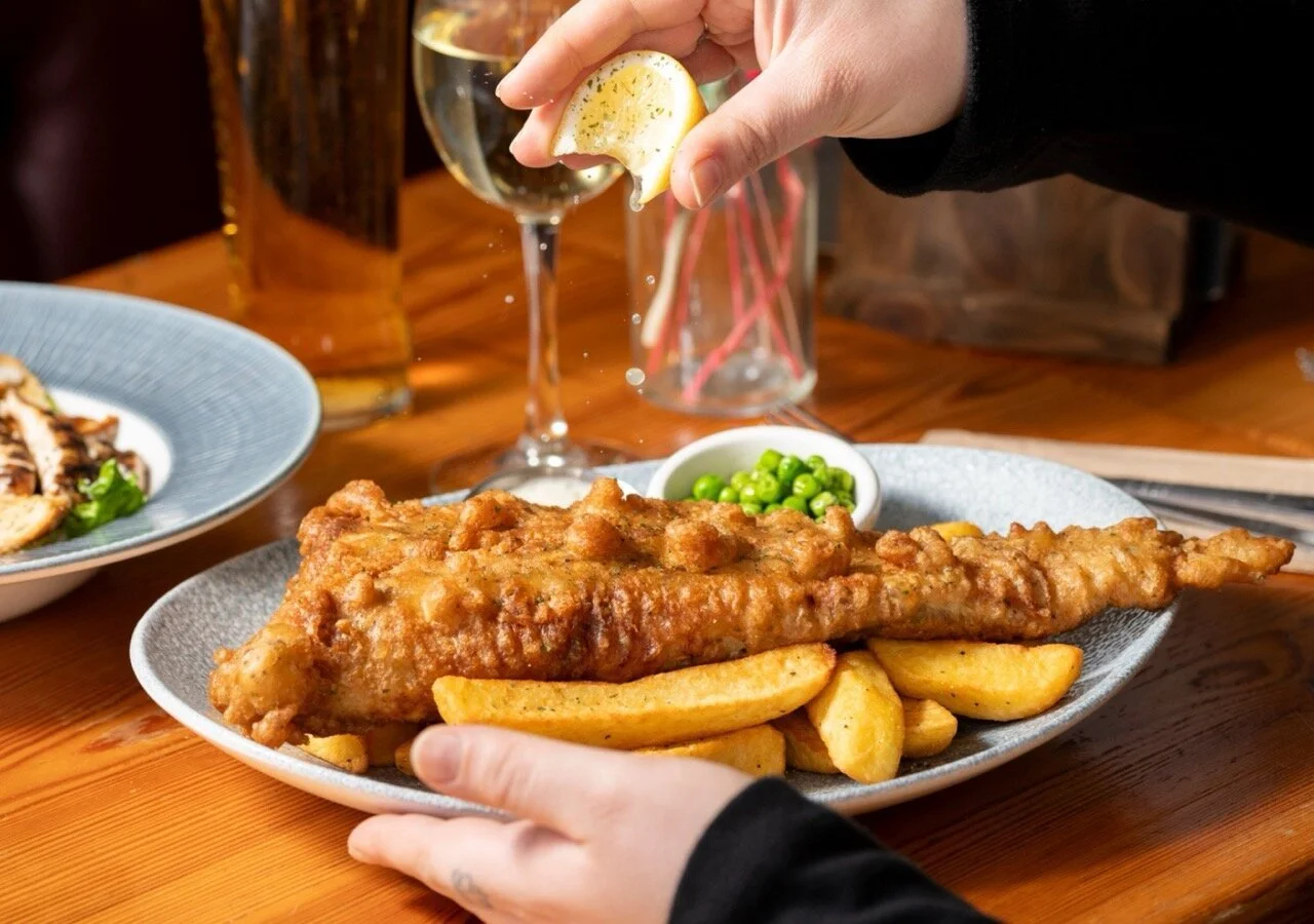 A person squeezing a lemon over a plate of fried fish and potato wedges with green peas inside a small bowl, on a wooden table with a glass of white wine in the background.