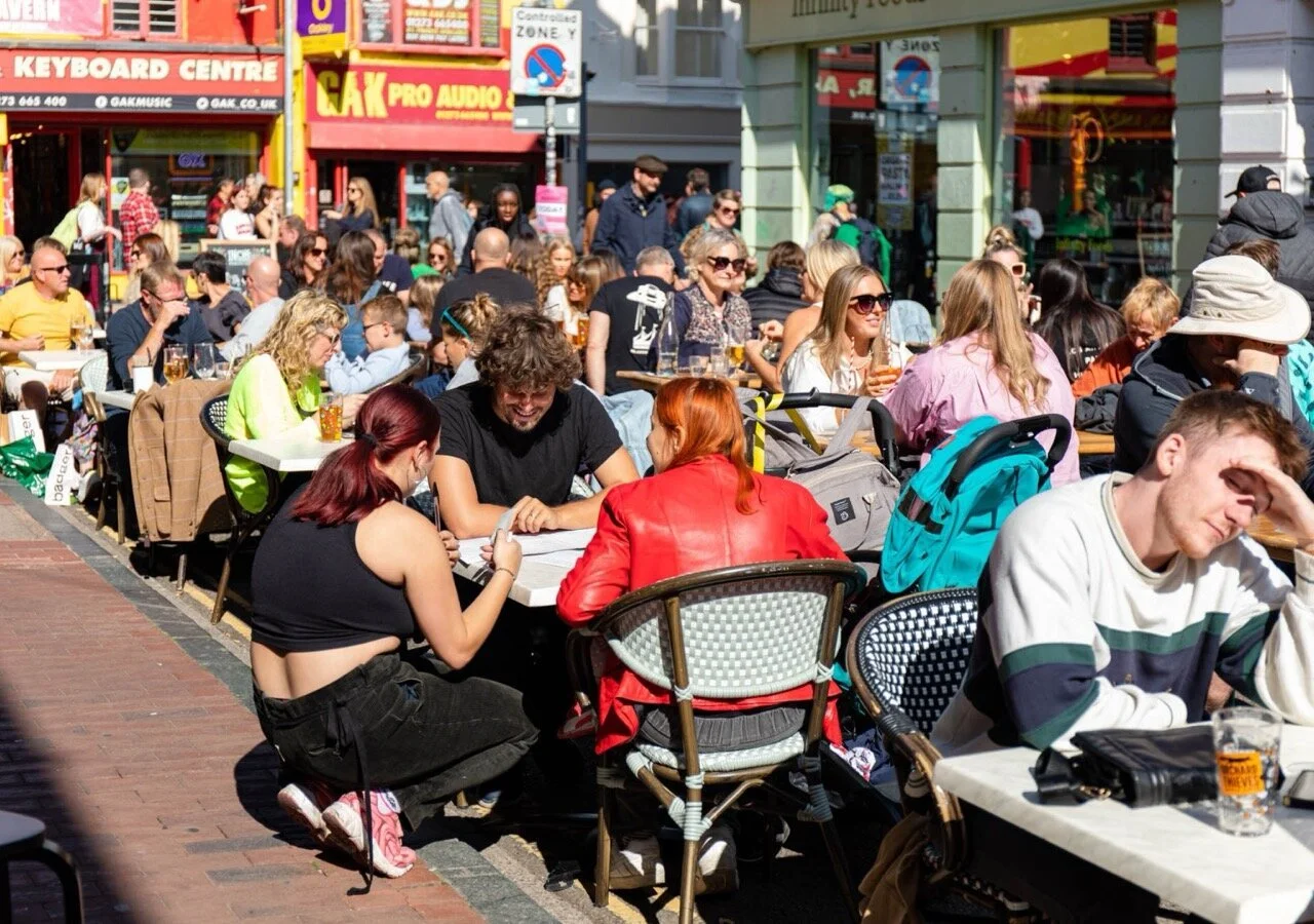 Crowded outdoor cafe with numerous tables and people enjoying sunny day, drinking and socializing on a city street.