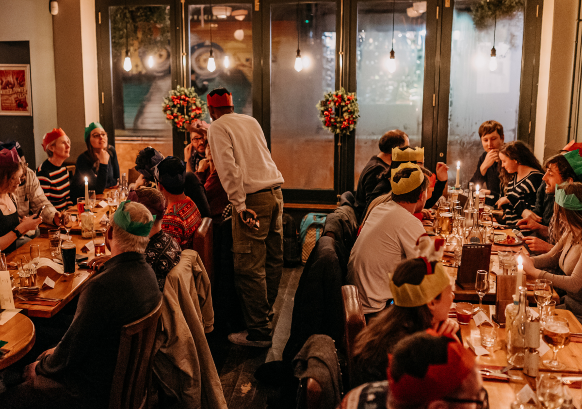 People gathered around a long dining table celebrating a holiday, wearing colorful paper crowns and festive hats, with dim lighting and holiday decorations.