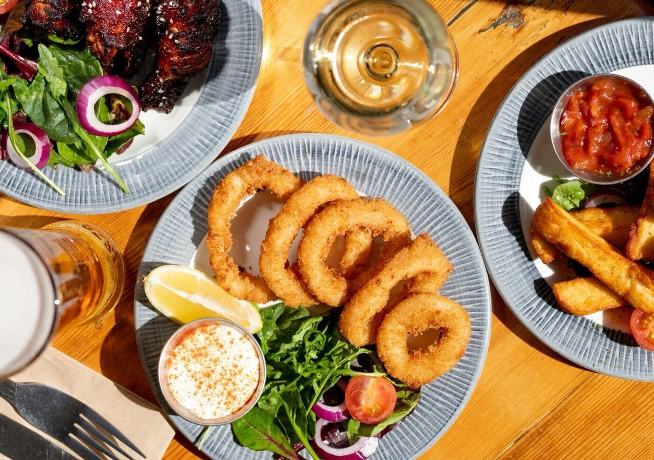 Fried onion rings with lemon wedge, salad, and dipping sauce on a blue plate, surrounded by additional salads, drinks, and food dishes on a wooden table.