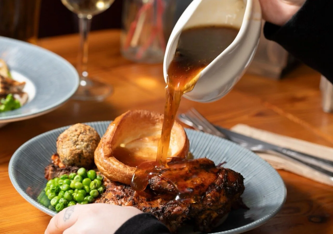 A person pouring gravy over a serving of carved beef with peas, stuffing ball, and a Yorkshire pudding on a blue plate, with wine glasses in the background.