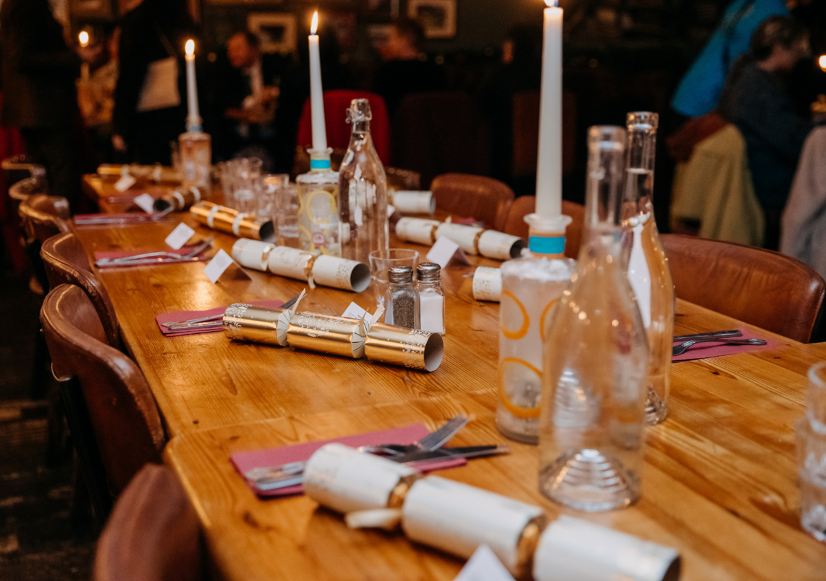 A long wooden dining table set for a celebration with candles, bottles, and rolled-up scrolls as decorations, in a dimly lit room filled with people.