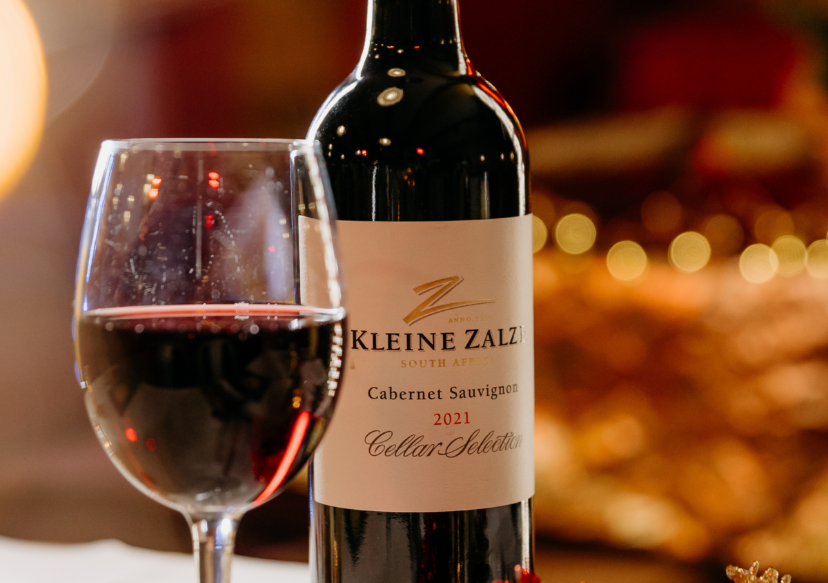A glass of red wine and a bottle of Kleine Zalze Cabernet Sauvignon from South Africa, vintage 2021, on a table with a warm, blurred background.