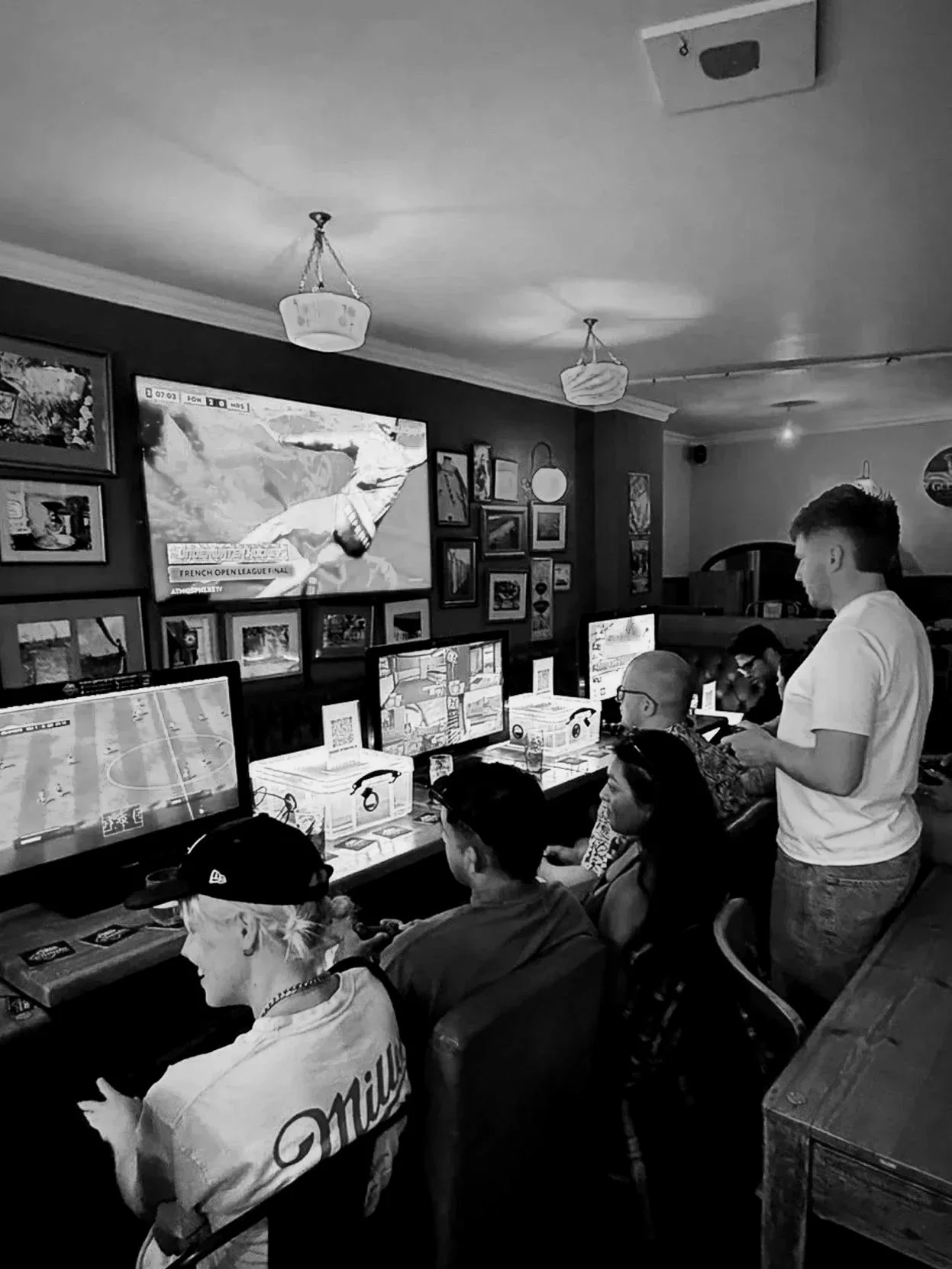 People playing video games on arcade machines in a gaming arcade, with a man standing and talking to a seated person, and pictures and a TV screen on the wall.