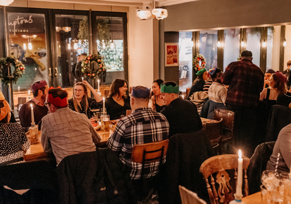 People sitting at a long wooden table in a cozy restaurant decorated with Christmas wreaths and candles, celebrating a holiday gathering.