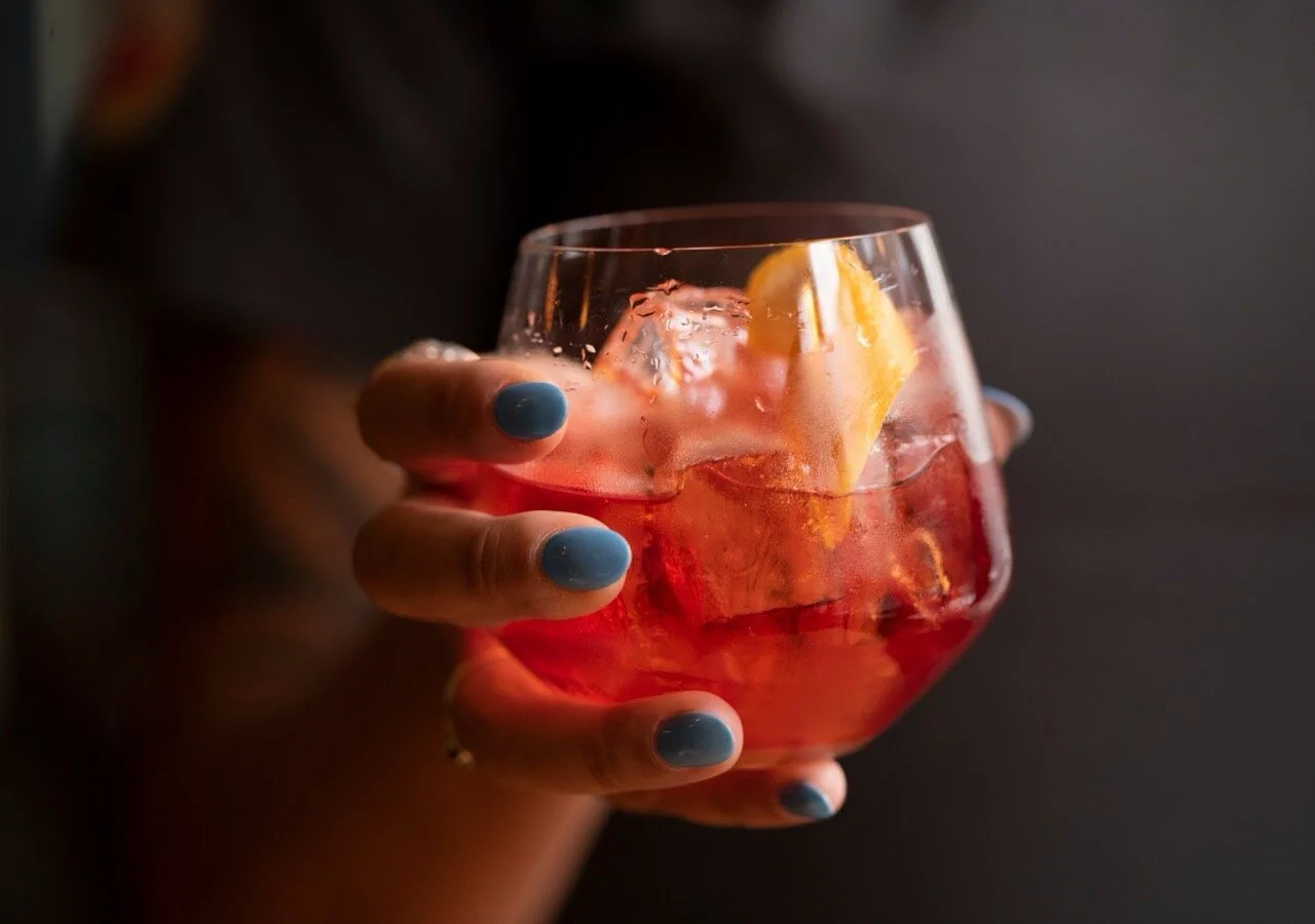 A person with blue painted nails holding a glass of pink drinks with ice and lemon wedge