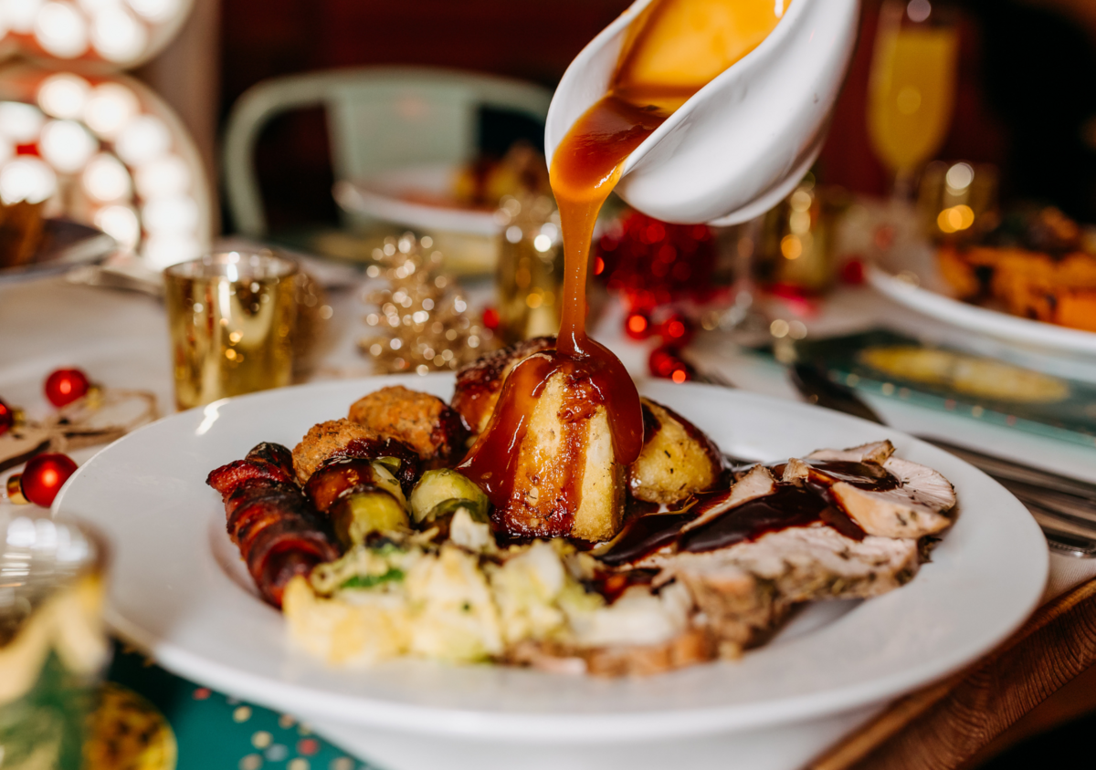 A holiday dinner plate with gravy being poured over a large piece of roast beef, with sides like mashed potatoes, stuffing, and vegetables, on a decorated table.