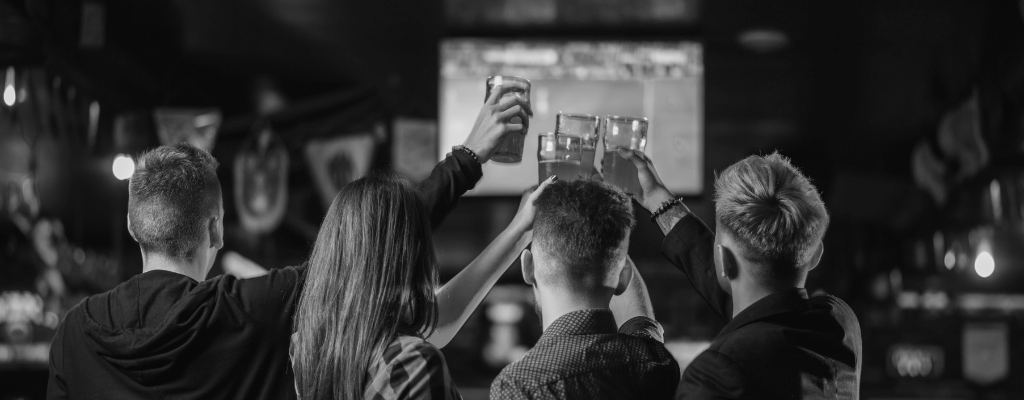Four people sitting at a bar raising their glasses for a toast, with a TV screen and sports flags in the background, in black and white.