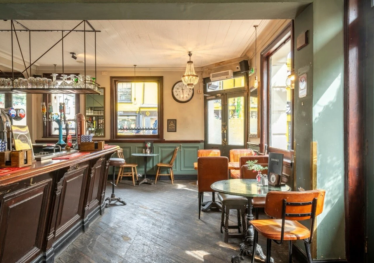 Interior of a cozy, vintage-style café or bar with wooden furniture, tables, and a bar counter. There are windows letting in natural light, a clock on the wall, and decorative lighting fixtures. The atmosphere appears warm and inviting.