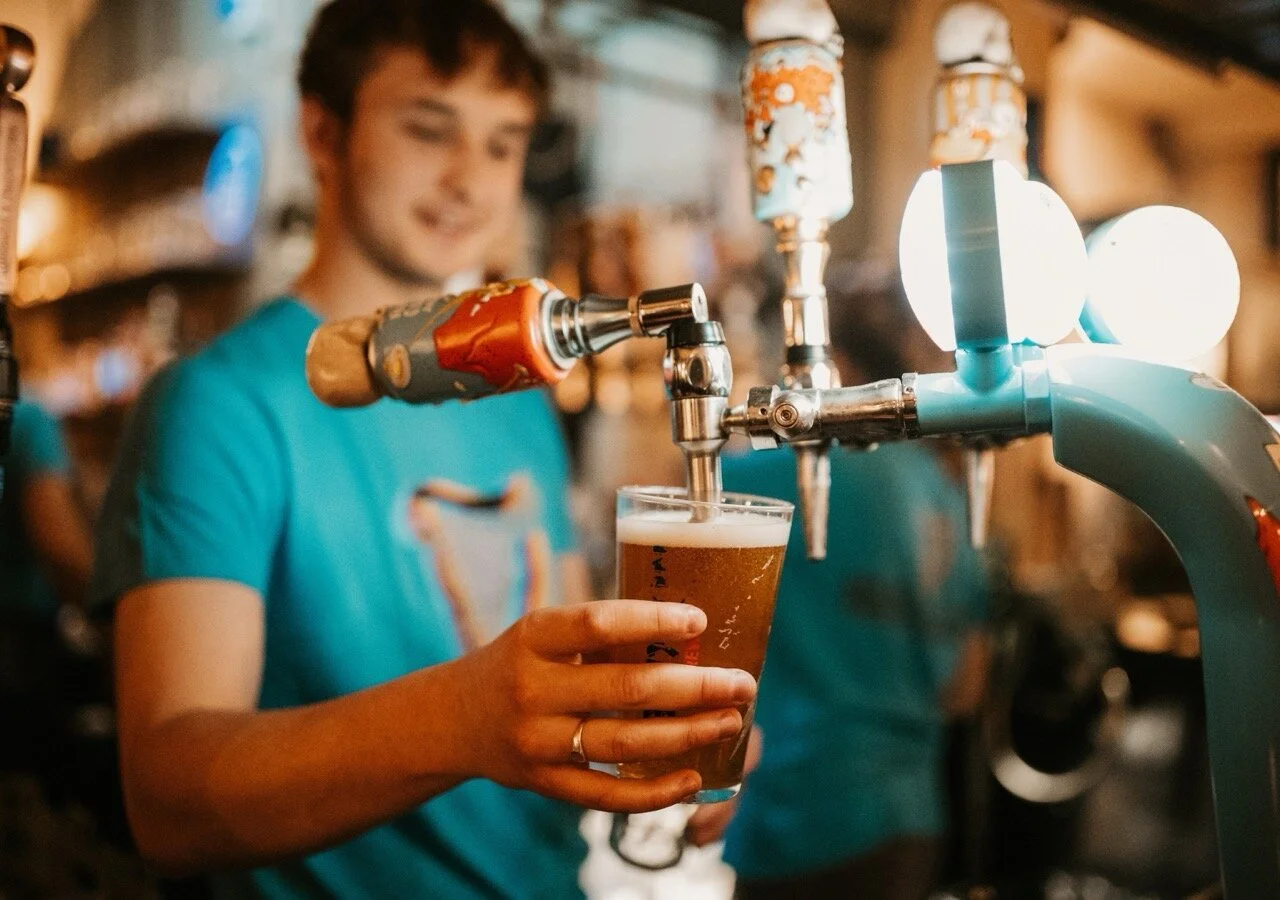 A person in a blue shirt pours beer from a tap into a glass in a bar or brewery setting.