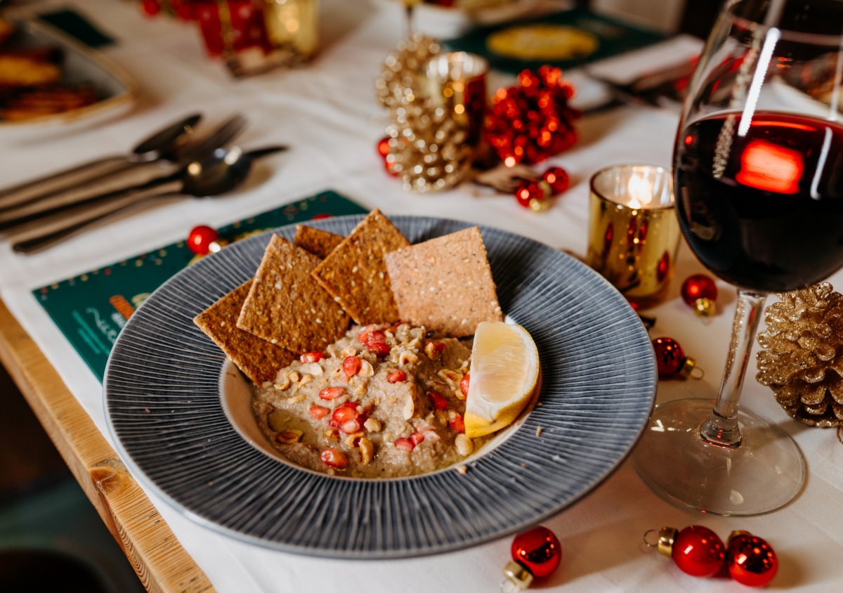 A festive holiday table setting featuring a plate of hummus topped with pomegranate seeds and walnuts, served with pita chips and lemon wedge. A glass of red wine and gold and red holiday decorations such as pinecones and ornaments are also on the ta