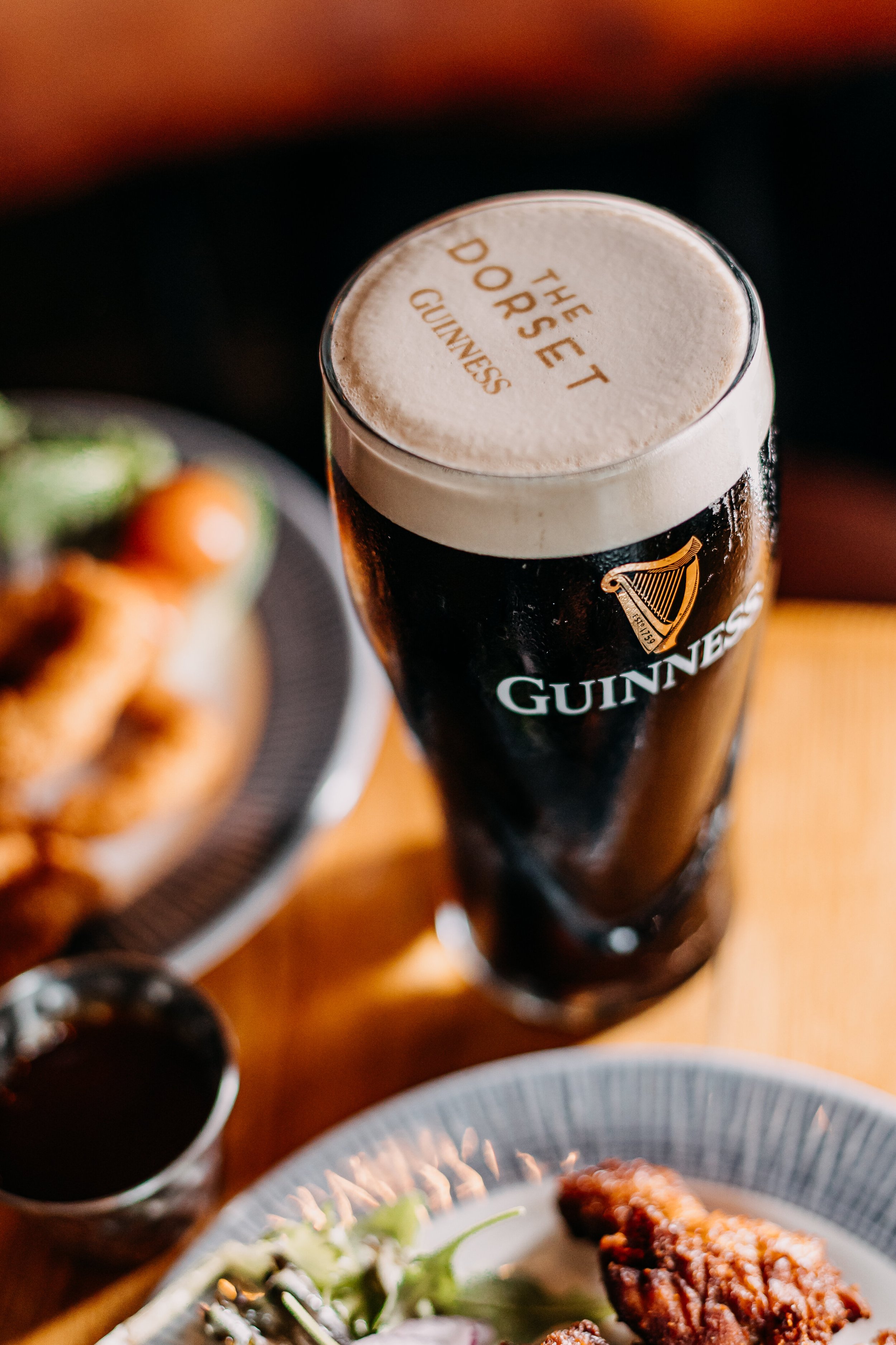 A pint of Guinness beer topped with a frothy head that has the words "The Do Set Do Rise Guinness" written on it, with food dishes and small containers of sauce in the background.