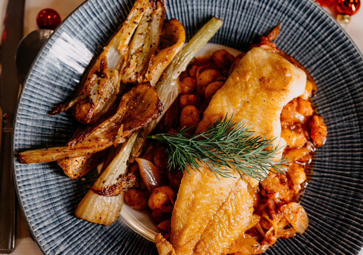 A plate with roasted vegetables, including carrots and onions, a baked fish fillet topped with a sprig of dill, and a side of roasted or grilled potatoes.