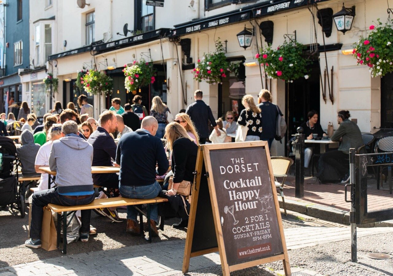 People dining at outdoor tables outside a restaurant called The Dorset, with a chalkboard sign advertising cocktail happy hour from 5 to 8 pm on Sundays through Fridays.