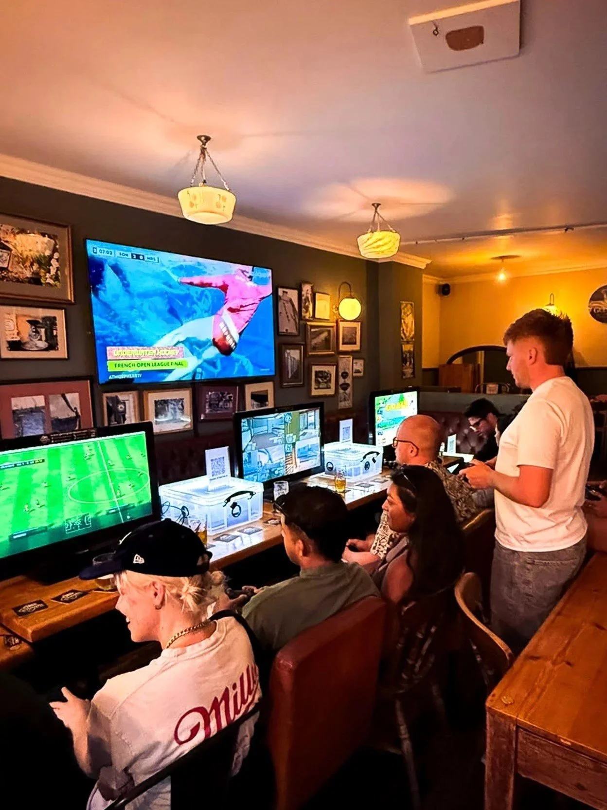 Group of people playing video games in a bar or gaming lounge, with multiple monitors and arcade machines, and a large TV displaying a sports event.