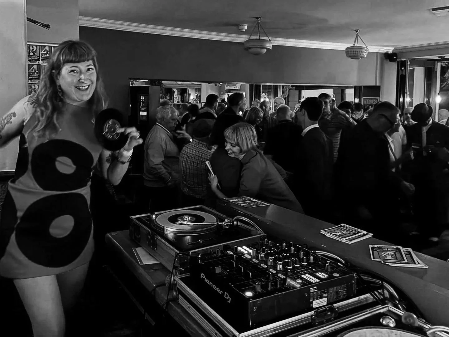 A woman smiling while holding a vinyl record in a crowded bar or club setting, with a DJ turntable and mixer in the foreground and a group of people socializing in the background.