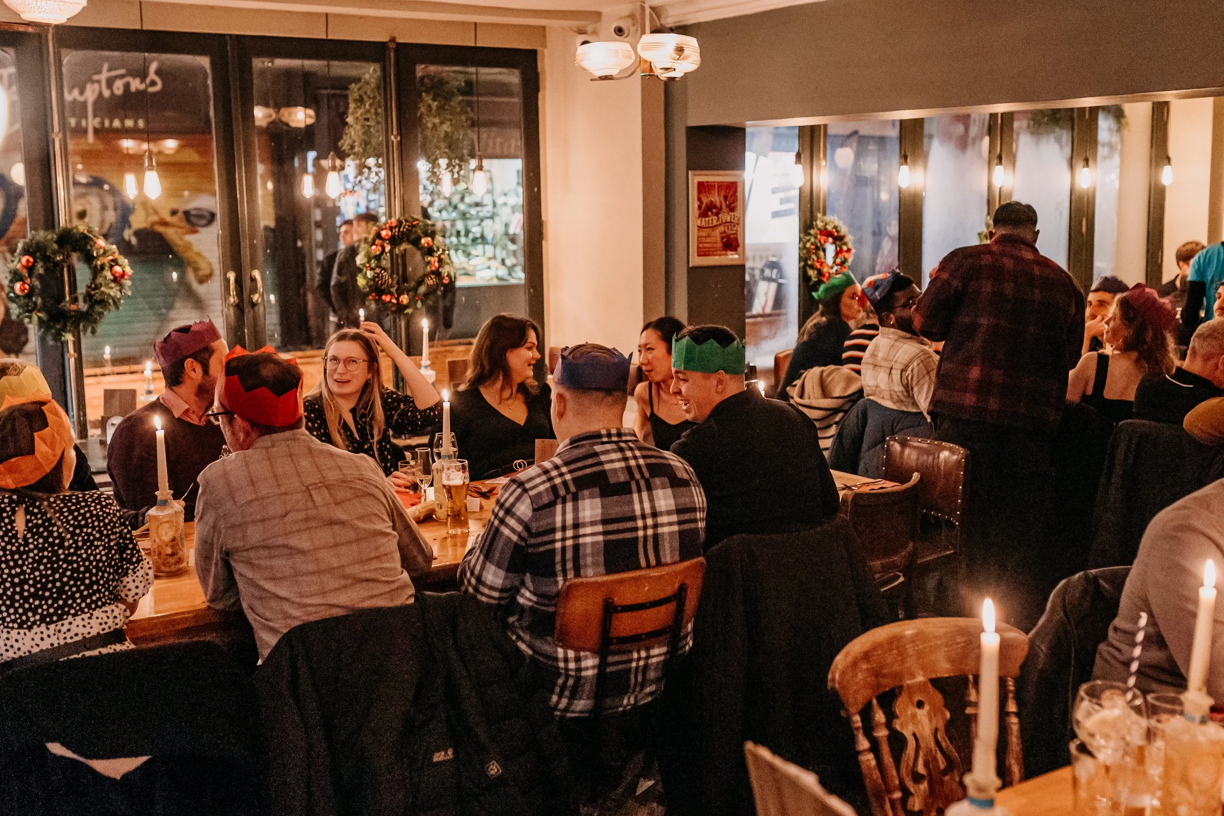 People gathered in a restaurant decorated for Christmas, wearing festive hats and scarves, with some smiling and chatting, seated around candles and enjoying drinks.