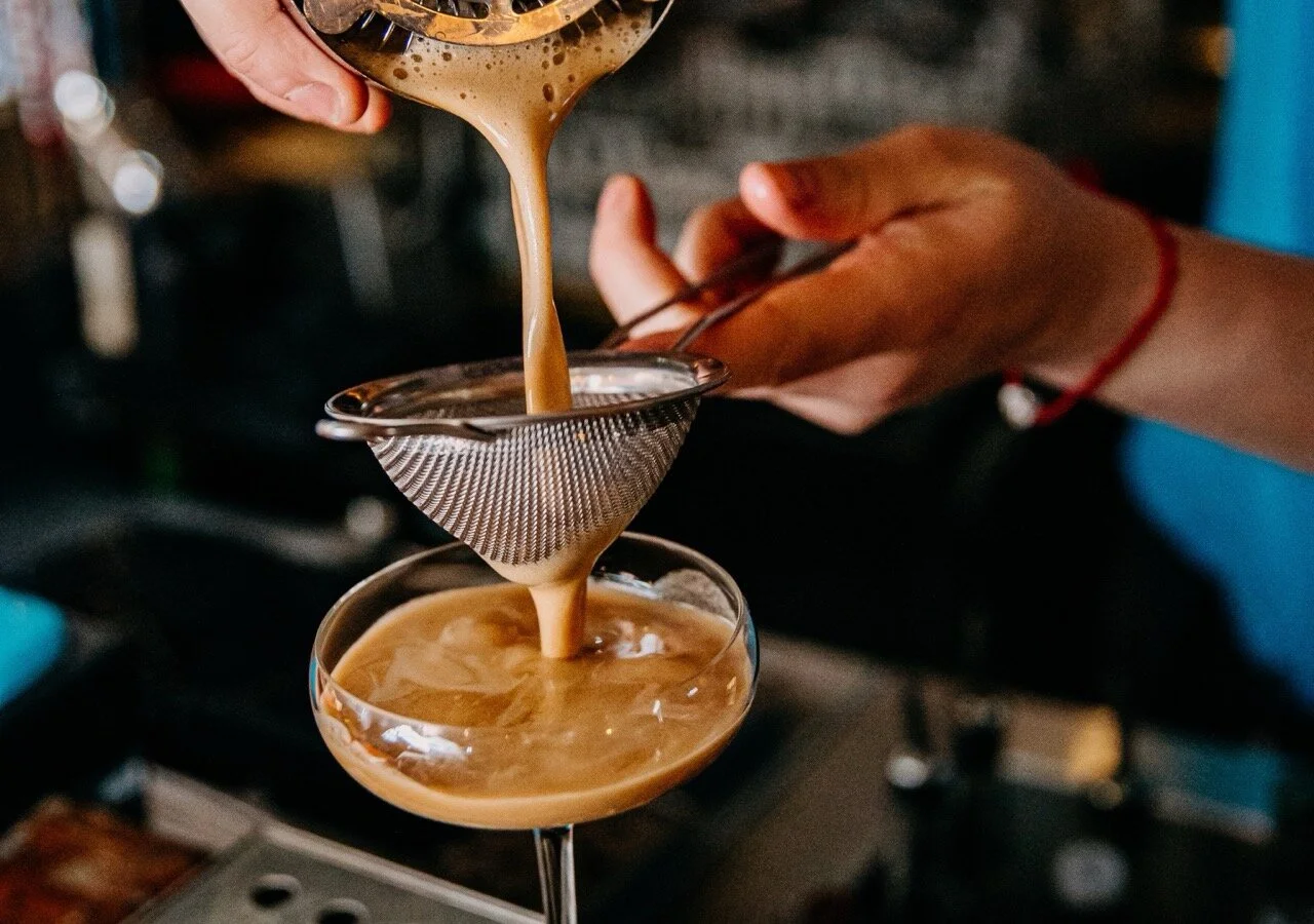 A person pours a creamy, light brown coffee beverage through a fine mesh strainer into a coupe glass, creating a layered drink.