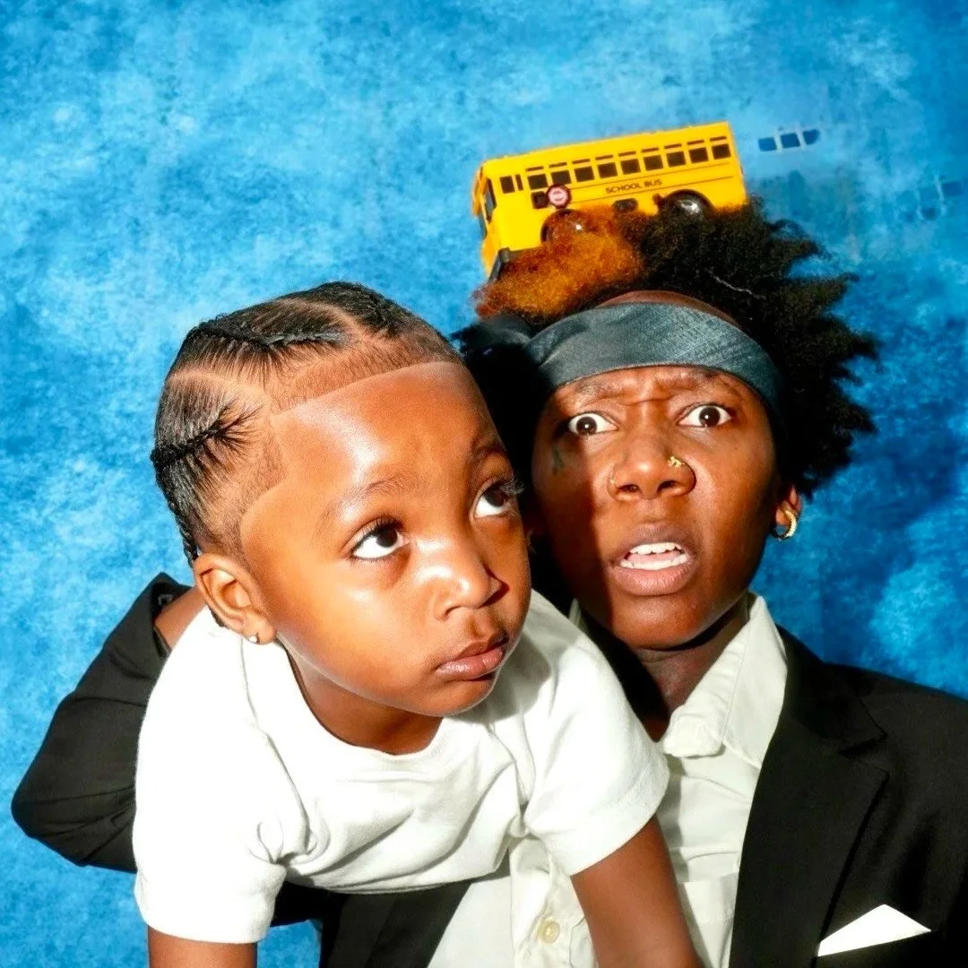 A woman lying on the floor with a yellow school bus toy on her head, looking surprised, while a young girl sits on her back, looking up, against a blue background.