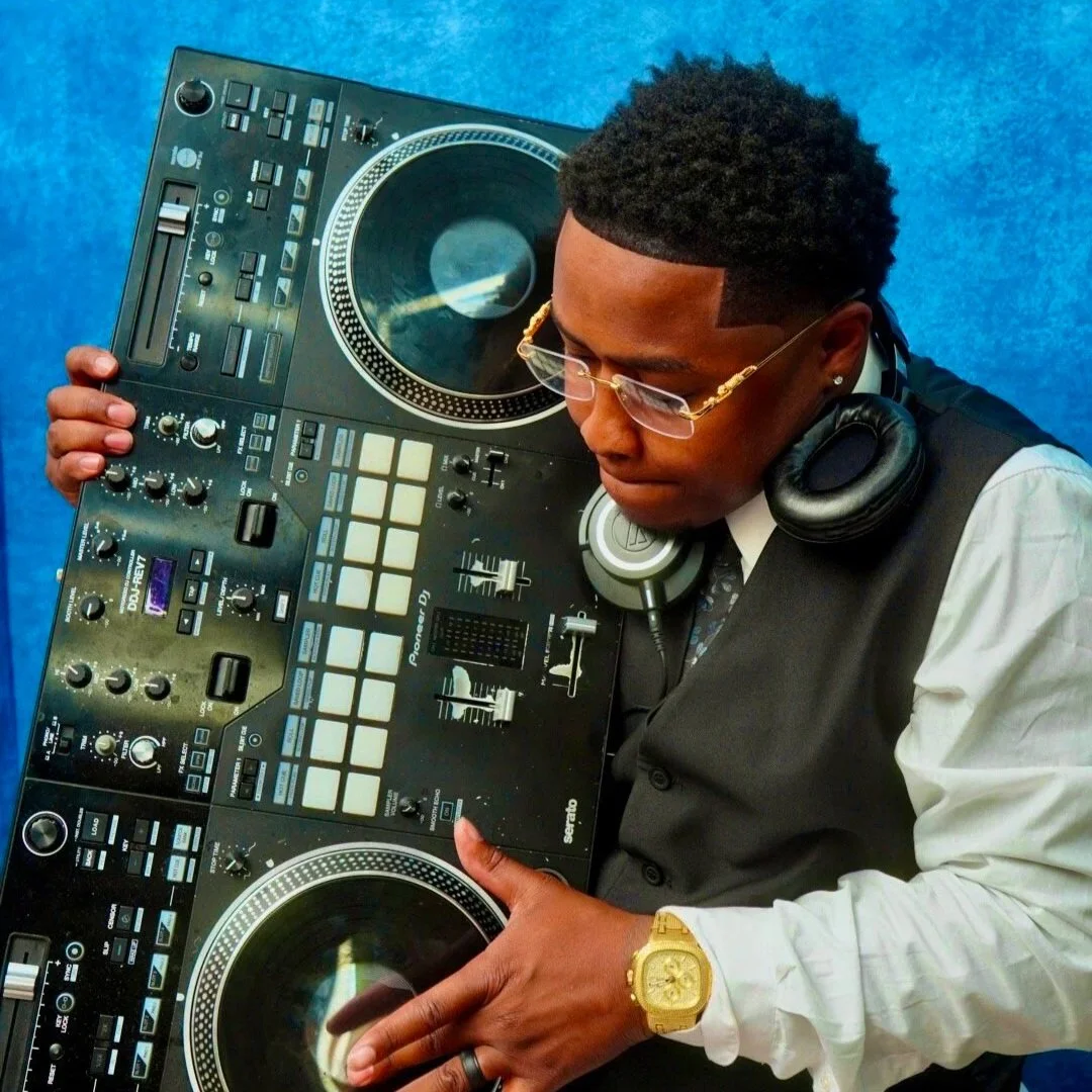 A DJ with short curly hair, wearing glasses, a white shirt, a black vest, and a gold watch, holding and adjusting a large DJ controller with both hands, against a blue background.