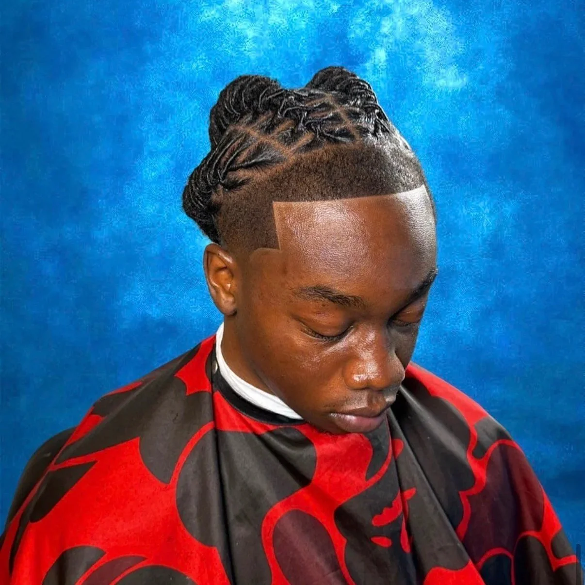 A person with a fresh, intricate hairstyle featuring braids and a high fade, seated against a blue background, wearing a red and black cape.