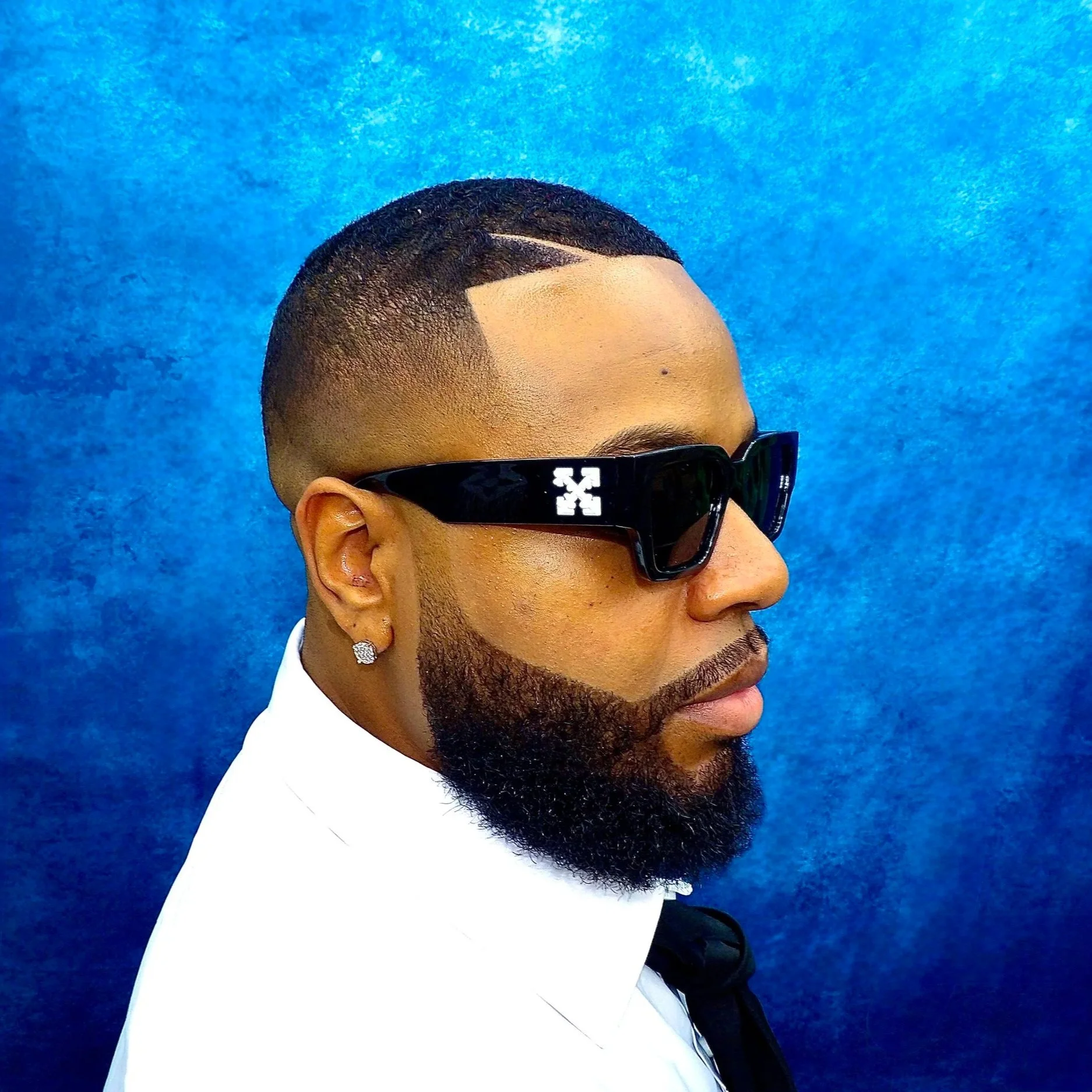 Close-up of a man with a neatly groomed beard, wearing black sunglasses and a white shirt, standing against a textured blue background.