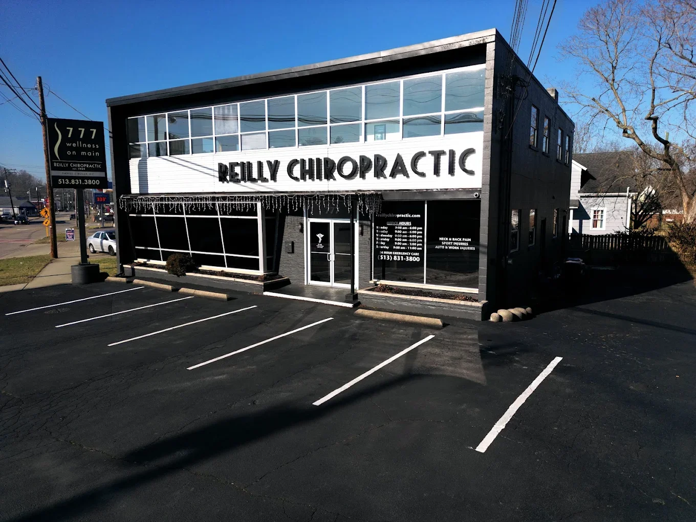 A two-story black and white building with the sign 'Reilly Chiropractic' on the front. There are parking spaces in front, some small bushes, and a street sign nearby. The building appears to be a chiropractic clinic.