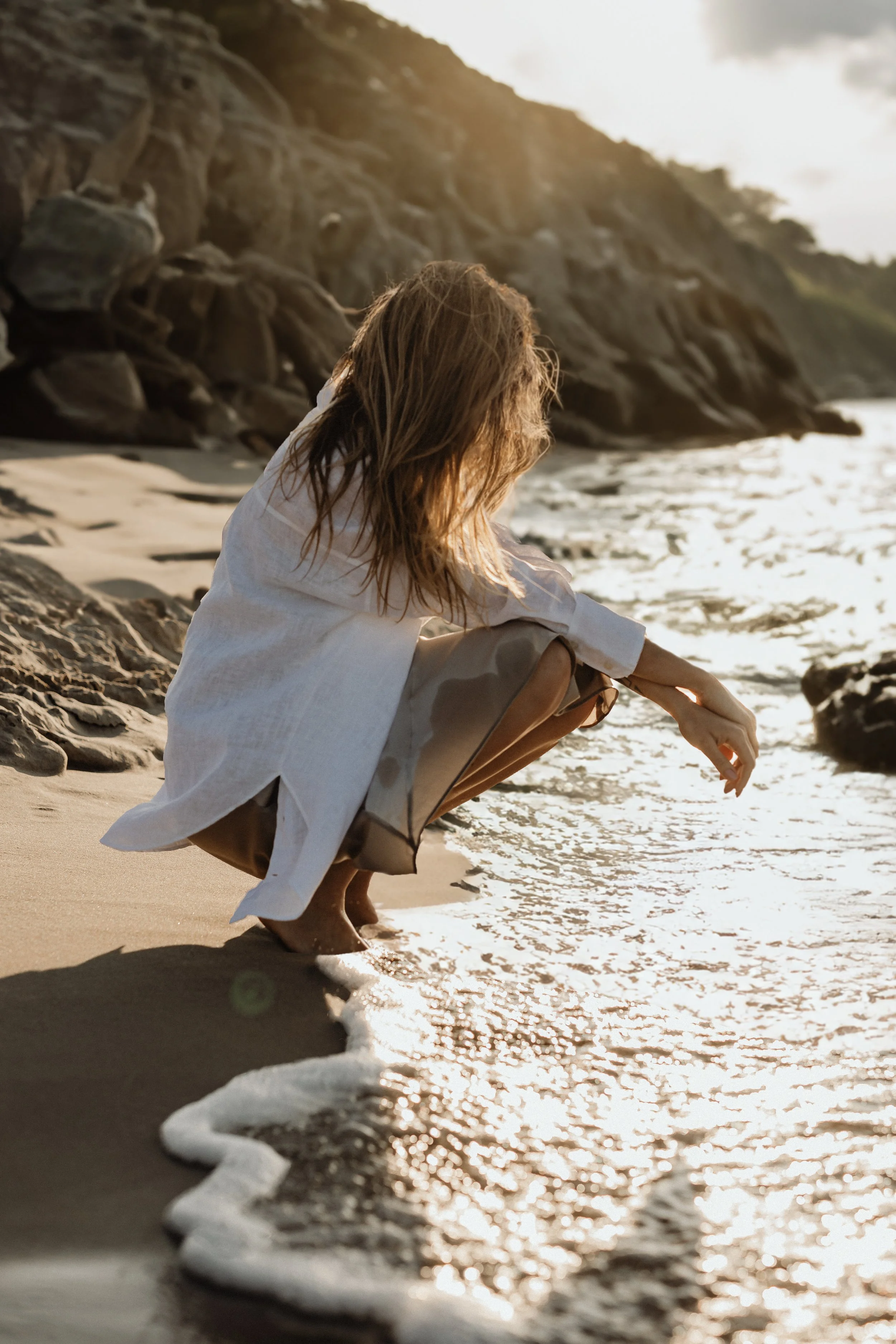 Person squatting on a sandy beach near the water, with rocky cliffs in the background and the sun setting or rising.