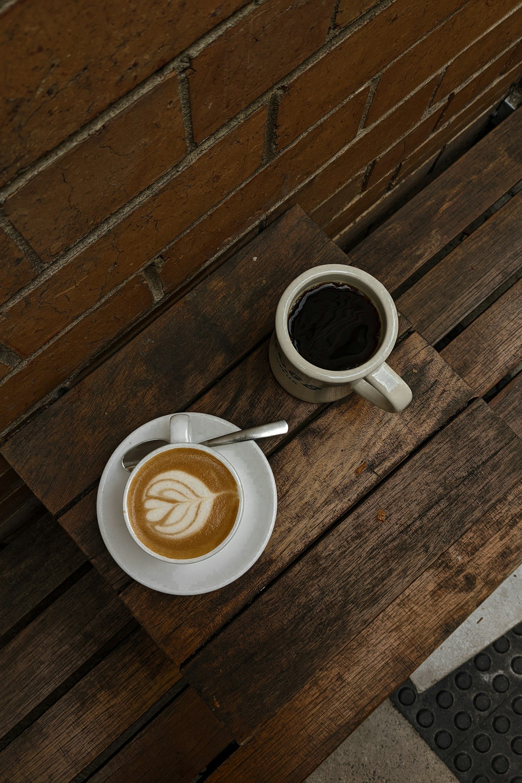 A white cup of coffee with latte art, a white cup of black coffee, and a small plate with a spoon on a rustic wooden table.