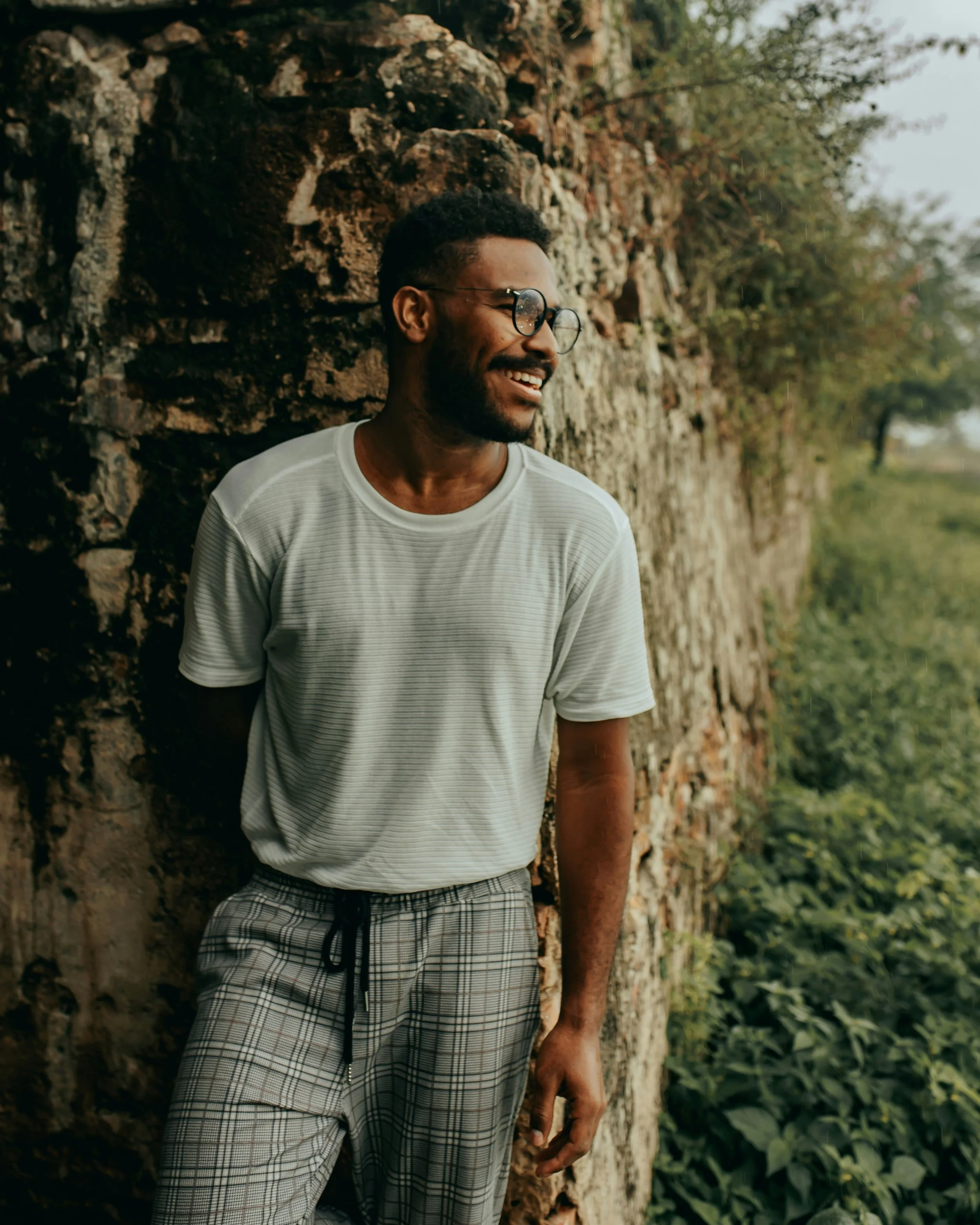 A smiling man with glasses leaning against a tree trunk outdoors in a natural setting with greenery.