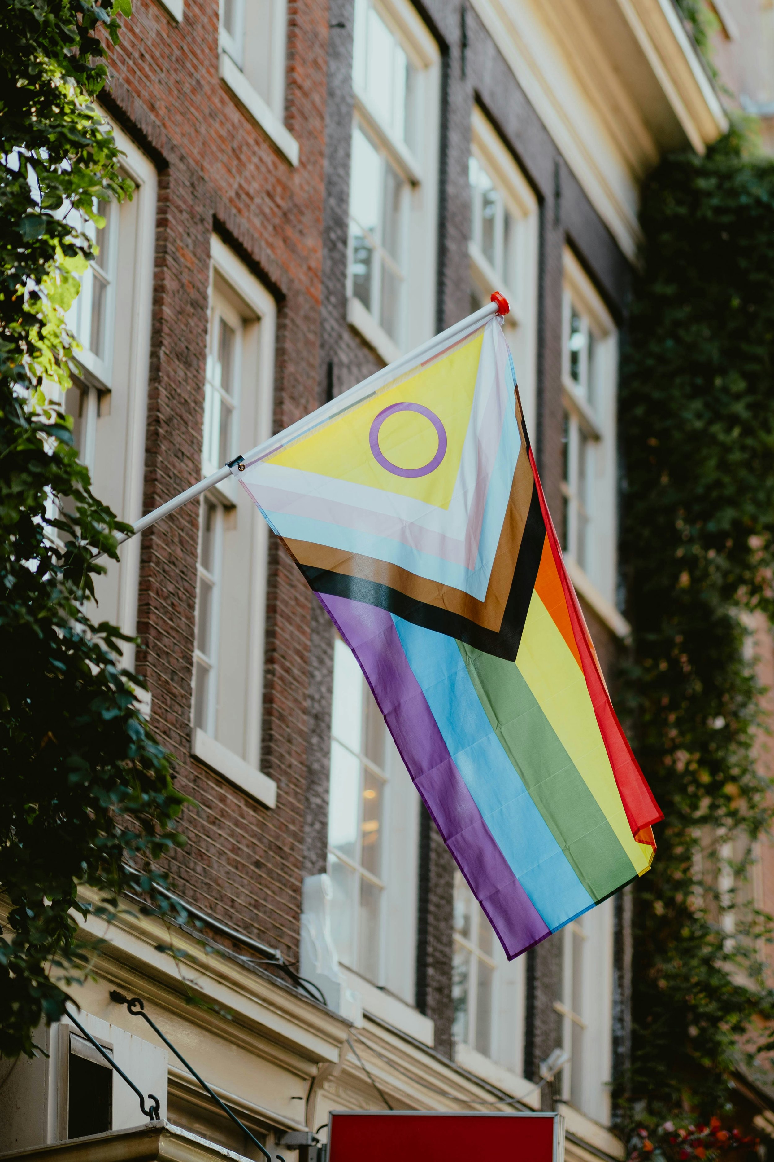 A rainbow pride flag with a yellow triangle and a purple circle hangs outside a brick apartment building, with trees and windows in the background.