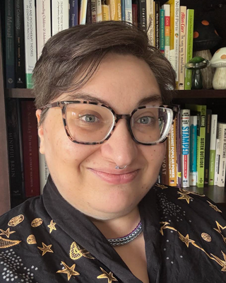 A woman with short brown hair, glasses, and a septum piercing taking a selfie in front of a bookshelf filled with books and decorative items.