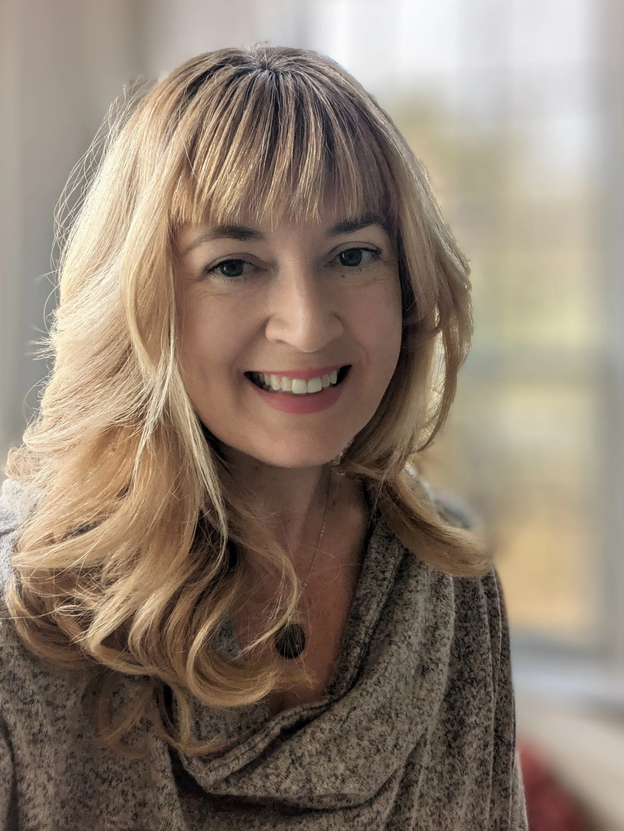 Close-up of a woman with long wavy blonde hair, smiling, wearing a grey sweater, indoors with blurred background.