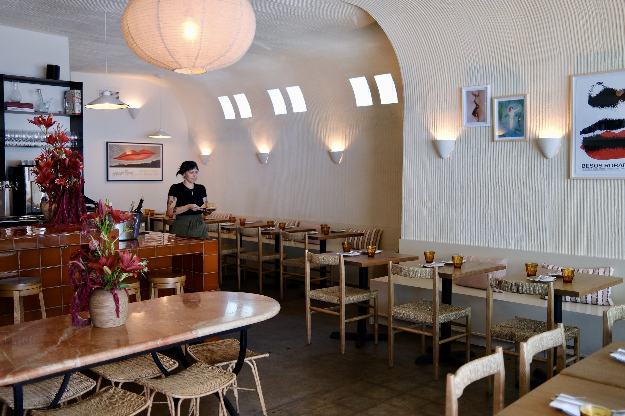Interior of a modern restaurant with a curved ceiling and large circular paper lantern light. There are framed art pieces on the wall, a woman serving food, and tables with glassware and flowers for decoration.