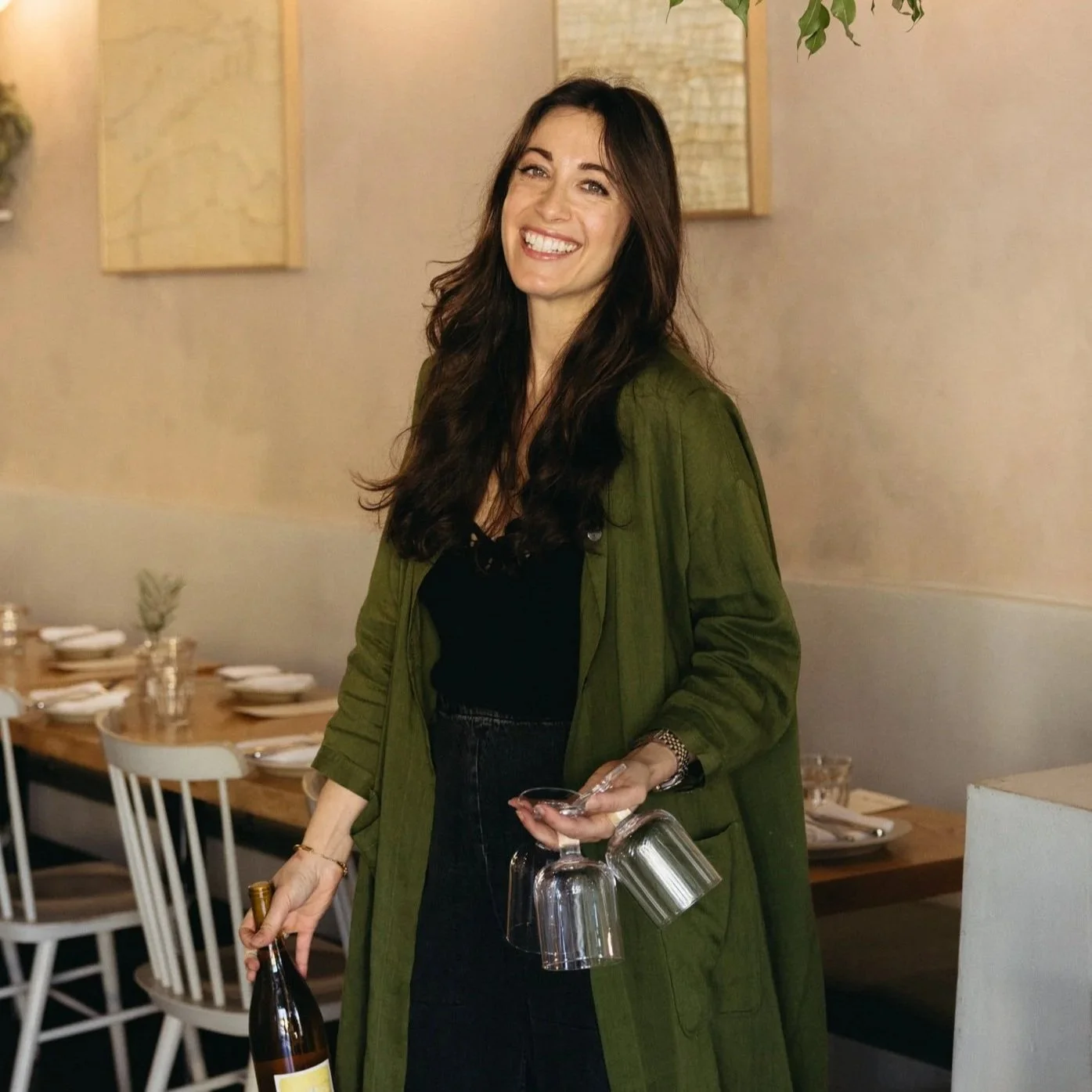 Smiling woman with long dark hair in a green coat, holding wine and upside-down wine glasses, in a dining setting with a long wooden table, chairs, and minimal decor