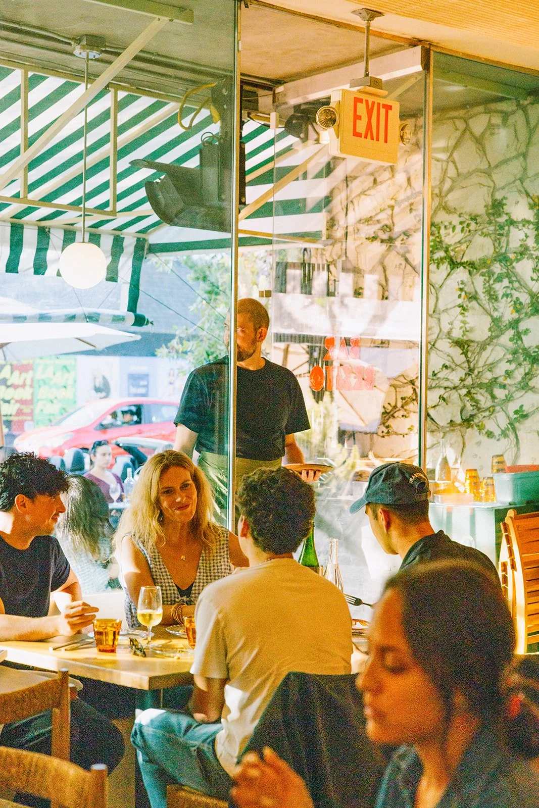 People dining inside a restaurant with large glass windows, a woman with blonde hair and a man wearing a baseball cap are visible, with a waiter serving food.