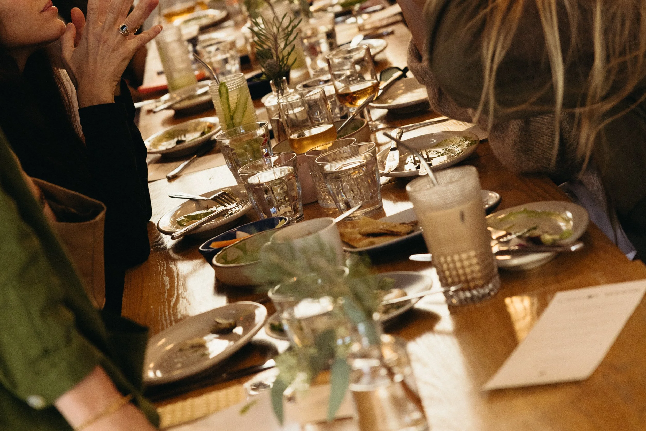 A group of people having dinner at a long wooden table with glasses, plates, and food in a warmly lit setting.