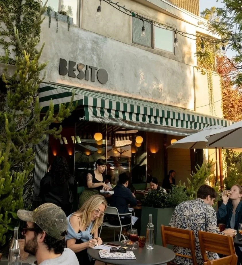 People dining outside a restaurant with a green and white striped awning in a cozy outdoor patio area, with string lights overhead and greenery around.