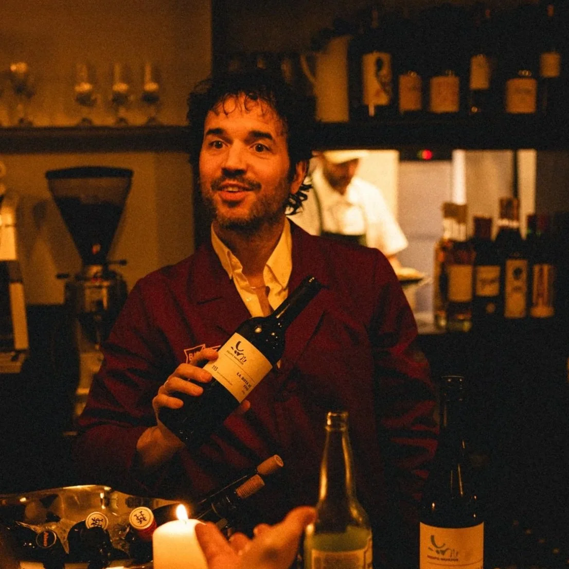 A man with dark curly hair and a beard smiling and holding a wine bottle in a dimly lit bar or restaurant. There are candles and bottles on the table and shelves behind him.