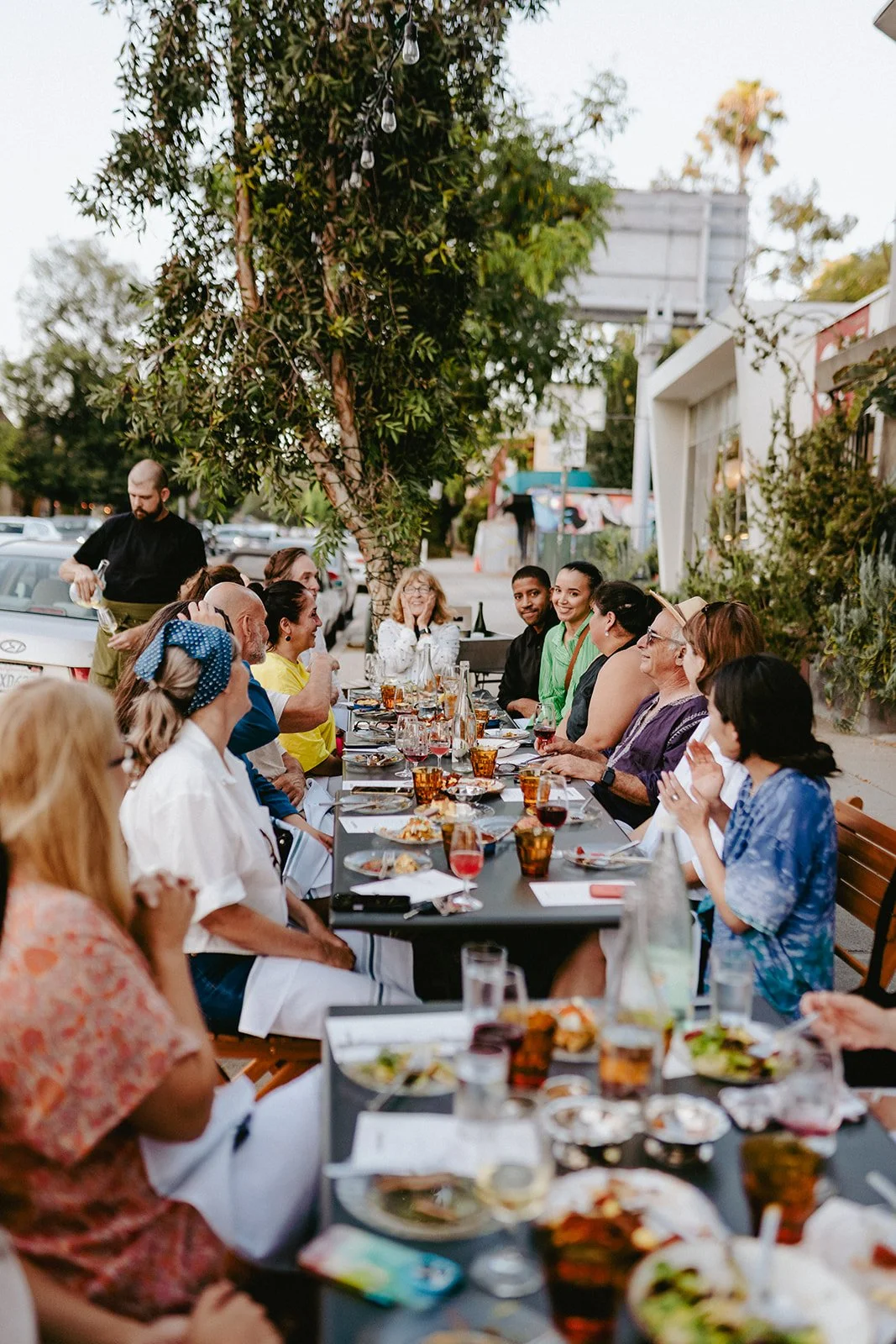 A group of people enjoying a meal together at an outdoor restaurant, sitting at a long table with food, drinks, and plates, under a tree with string lights.