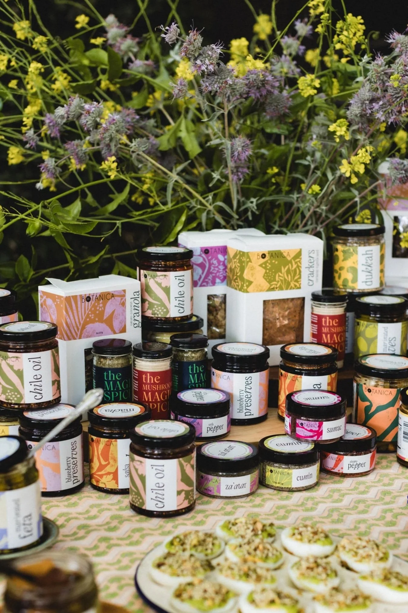Assorted jars of spices and seasonings with labels, placed on a table with a backdrop of purple, yellow, and green flowers, and plates of prepared food such as deviled eggs.