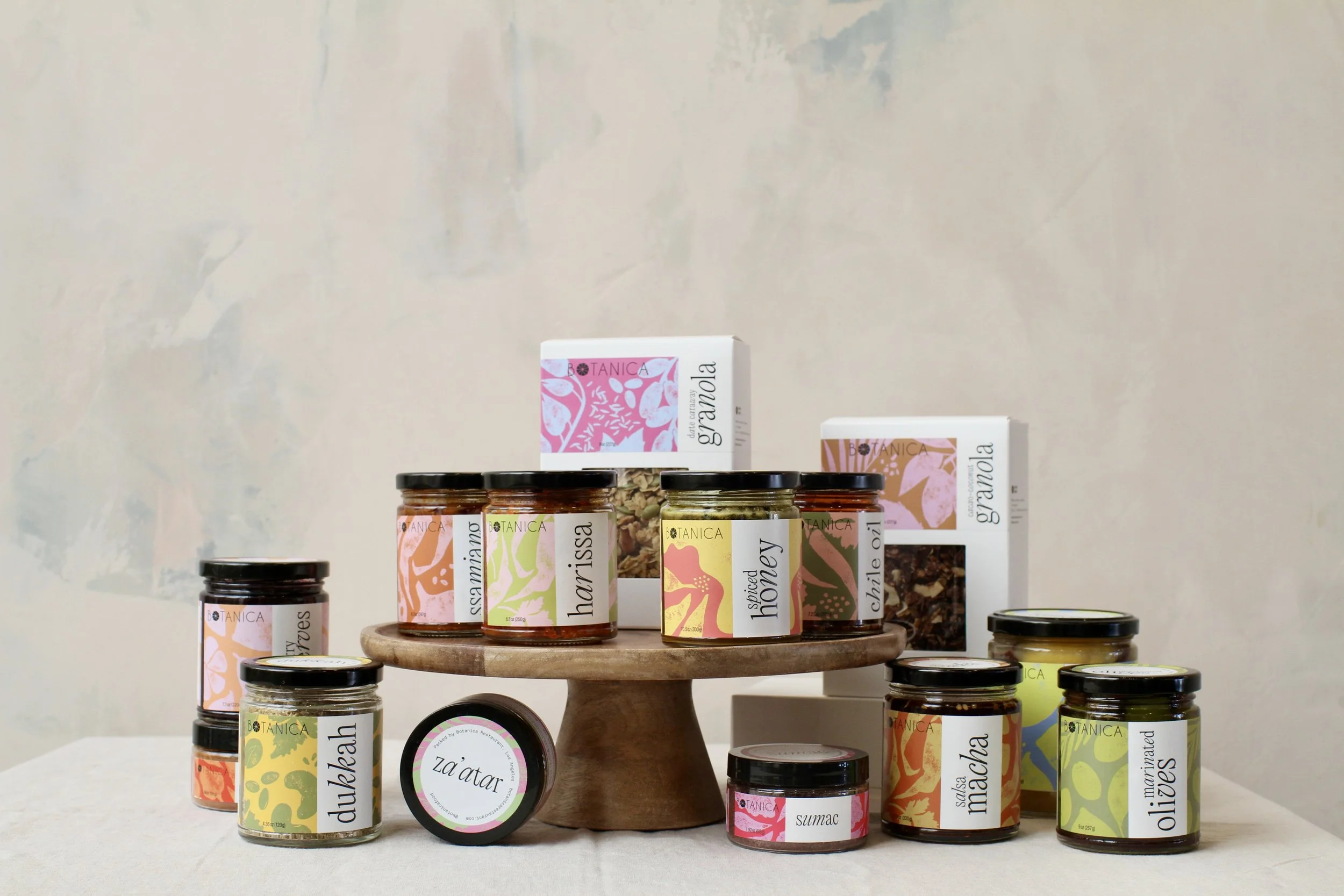 Assorted jars of organic food products arranged on a round wooden display stand, with some boxes of products behind on a shelf against a light-colored wall.