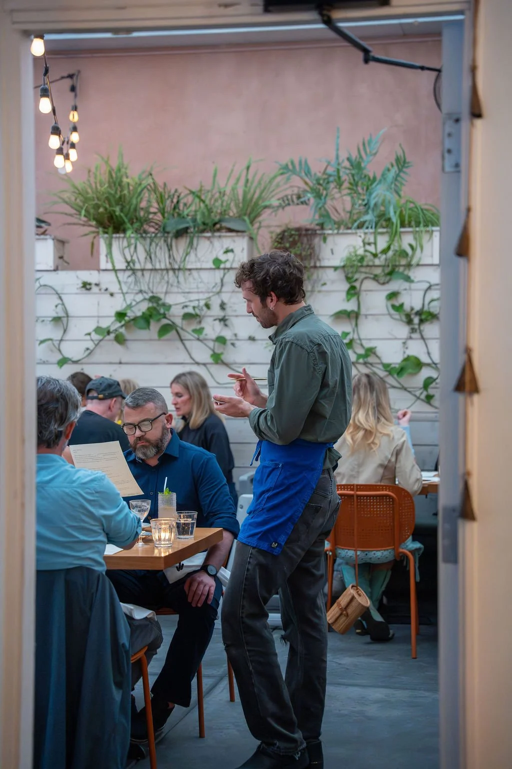 A waiter taking an order from a group of people dining at a restaurant with a wall decorated with green plants and warm lighting.