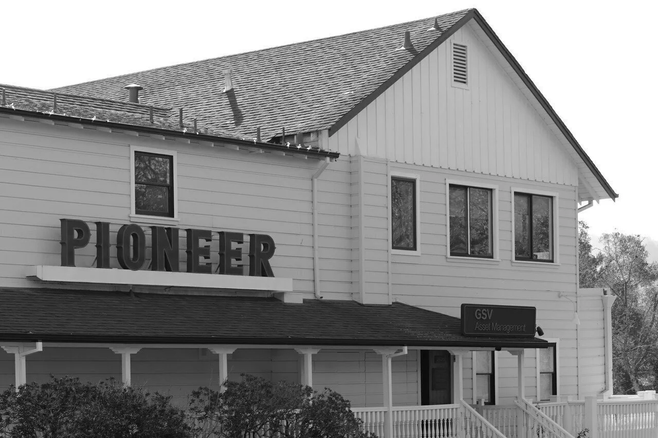 A two-story white building with a sign that says 'PIONEER' and a smaller sign reading 'GSV Asset Management', featuring a porch with stairs, windows, and a sloped roof.