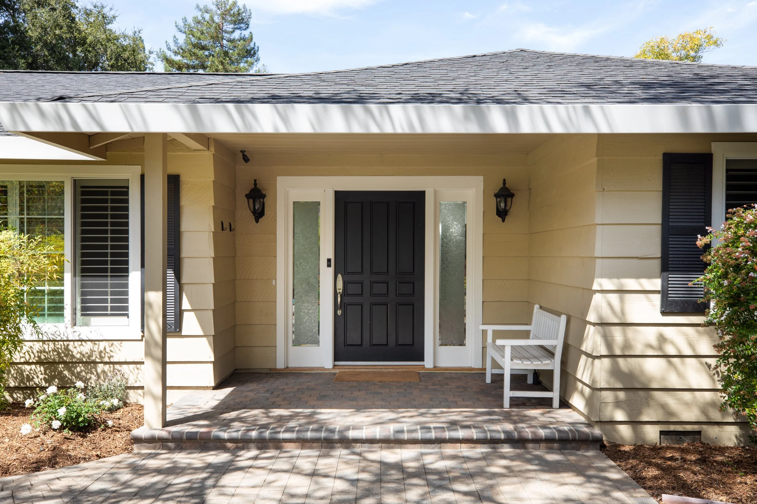 Front porch of a house with a black door, two black wall-mounted lamps, a white bench, and a brick-paved pathway, with trees and shrubs in the background.